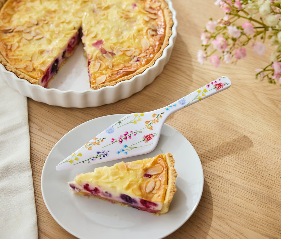 Obstkuchen mit Mandeln in einer weißen Form, ein Stück Kuchen auf einem Teller und ein Tortenheber mit Blumenmuster.