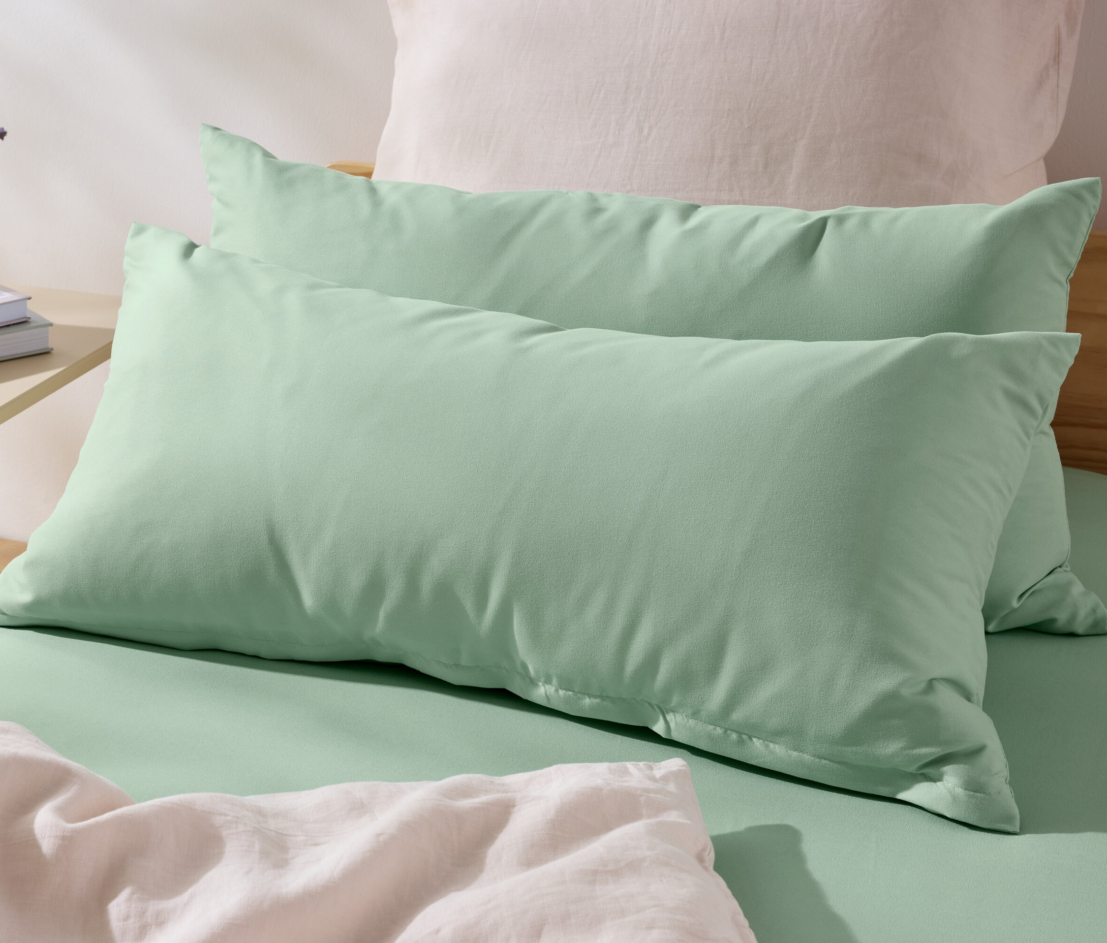 Zwei grüne Microfaser-Kopfkissenbezüge (ca. 80 x 40 cm) liegen auf einem Bett. Auf dem Nachttisch stehen eine Vase mit Blumen und Bücher.