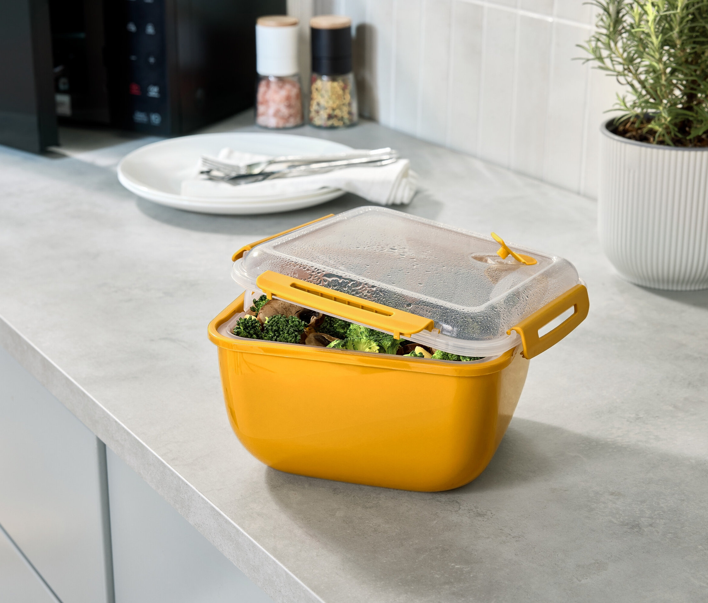 Gelbe Frischhaltedose mit Brokkoli und Pilzen unter einem transparenten Deckel auf einer Küchentheke.