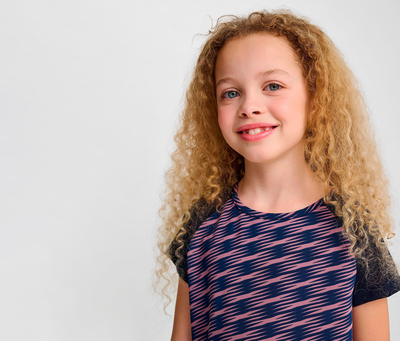 Porträt eines lächelnden Mädchens mit lockigen blonden Haaren in einem HUMMEL HMLJR Mix T-Shirt S/S.