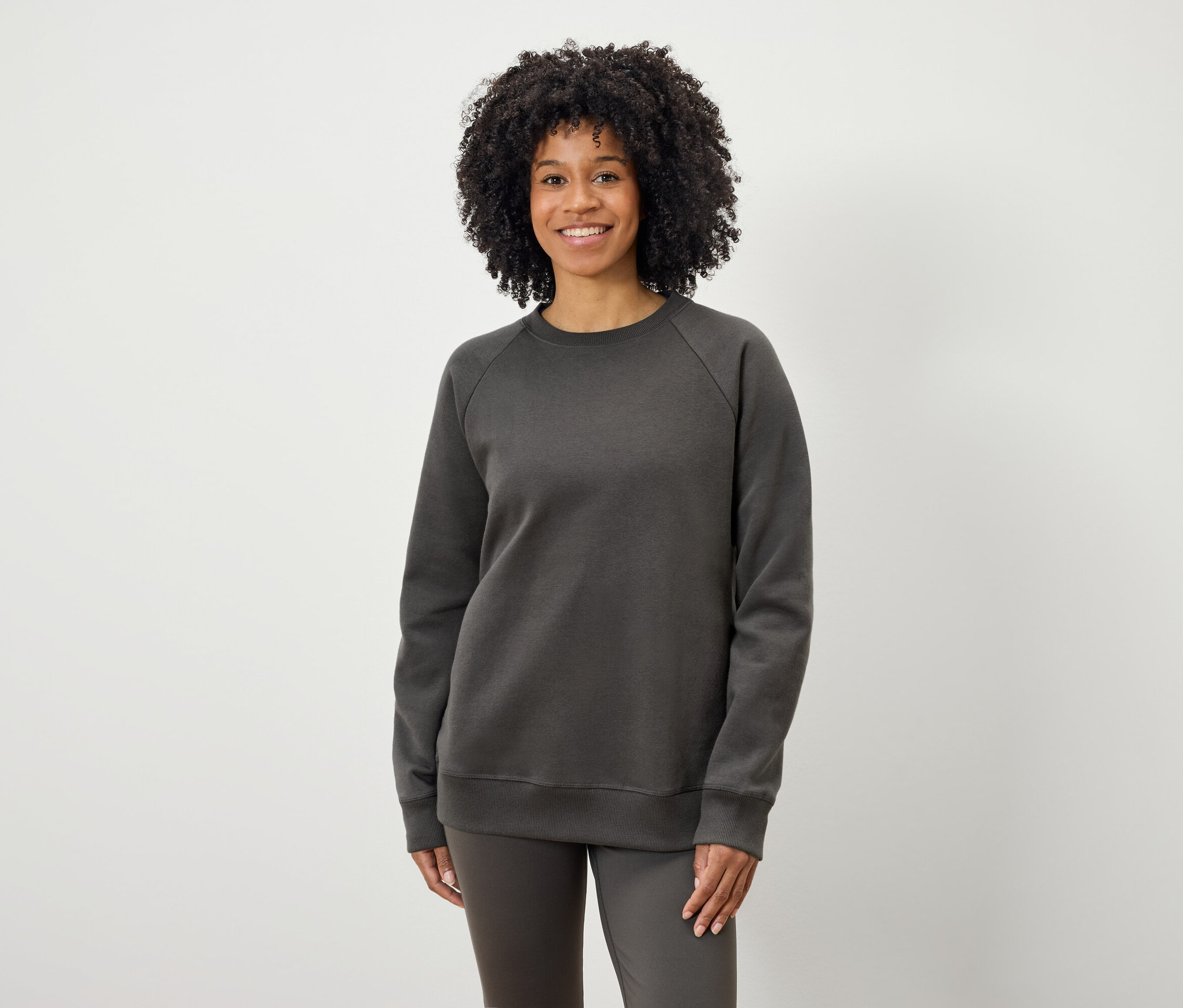 Eine Schwarze Frau mit lockigem Haar posiert vor einem hellen Hintergrund in einem grauen Sweatshirt und grauen Hosen.