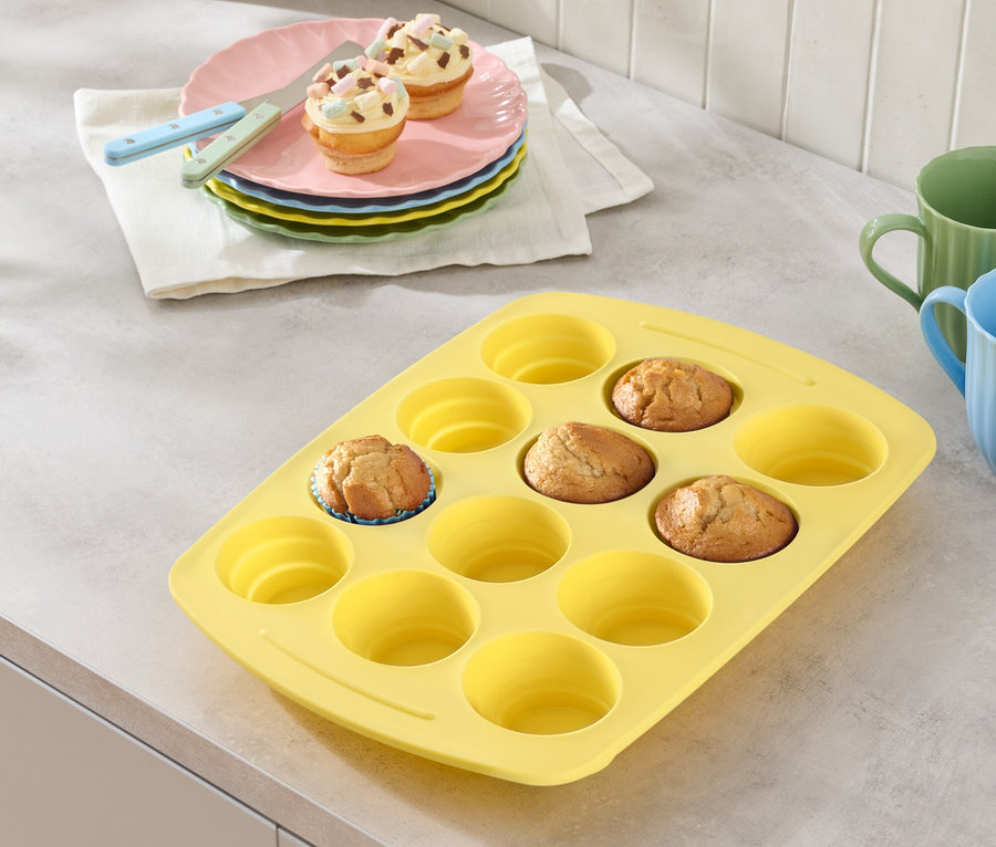 Gelbe Muffinform mit ein paar Muffins, Teller mit Muffins und Gläser auf einer Arbeitsfläche.