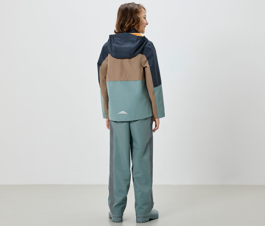 Mädchen von hinten in bunter Jacke und Kinder-Regenhose, salbei.