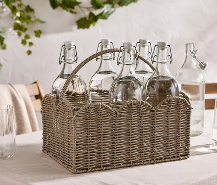 Sechs Wasserflaschen mit Bügelverschluss in einem Weidenkorb auf einem Tisch, dazu zwei Gläser.