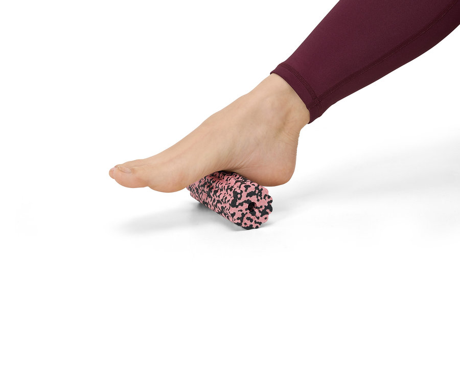 Fuß in Burgunder-Leggings rollt über eine rosa-schwarz gesprenkelte Faszienrolle auf weißem Grund.