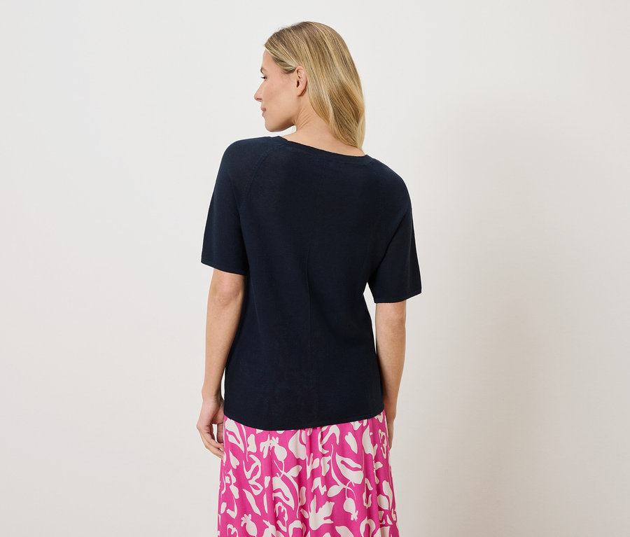 Eine Frau mit blonden Haaren trägt ein marineblaues T-Shirt und einen rosa-weiß gemusterten Rock, von hinten gesehen.