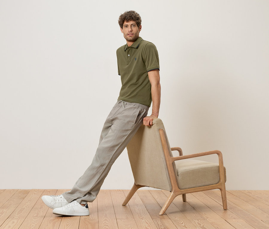 Ein Mann lehnt an einem beigen Sessel, trägt ein olivgrünes Poloshirt, graue Leinenhose und weiße Sneaker.