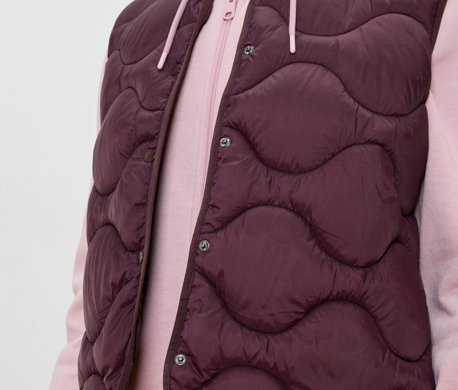 Person trägt eine ärmellose, auberginefarbene Steppweste über einem rosa Hoodie.