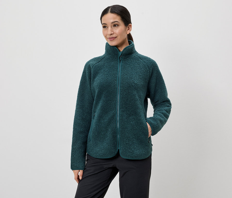 Frau mit Zopf trägt eine grüne Fleecejacke mit Reißverschluss und schwarze Hose, eine Hand in der Jackentasche.