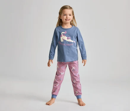 Ein junges Mädchen steht barfuß und lächelt in einem blauen Langarmshirt mit einer streckenden Katze und rosa Hosen mit Einhornmuster.