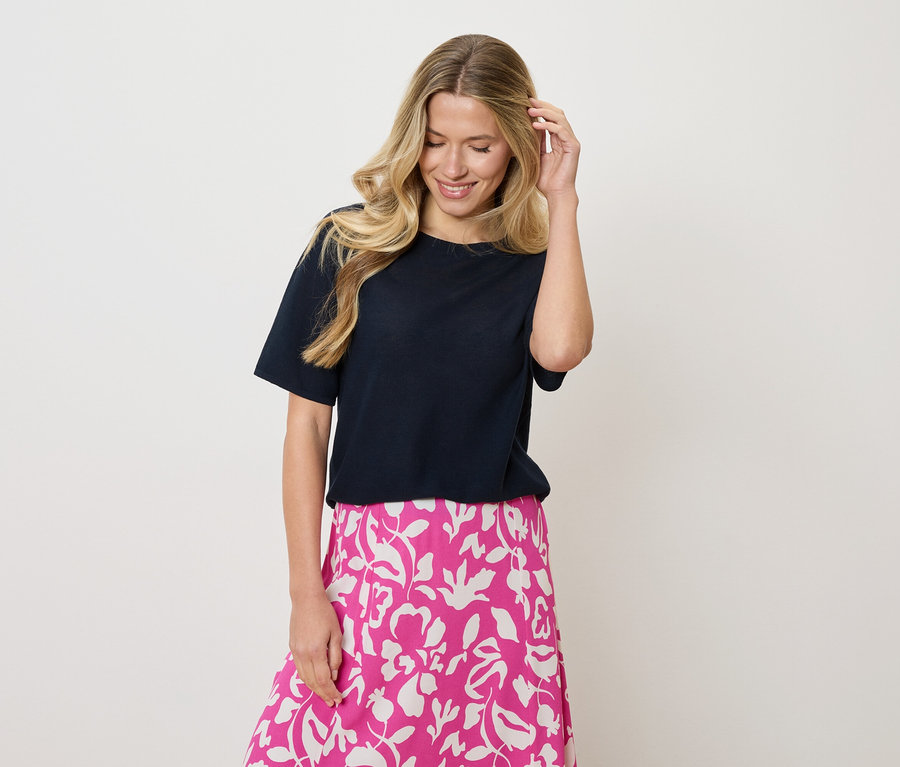 Frau mit blonden, gewellten Haaren, die ein marineblaues T-Shirt und einen rosa-weißen Rock mit Blumenmuster trägt und mit einer Hand ihr Haar berührt.