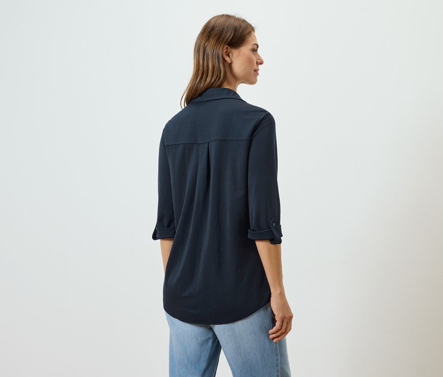 Rückansicht einer Frau mit langen braunen Haaren, die eine marineblaue Hemdbluse und hellblaue Jeans trägt.