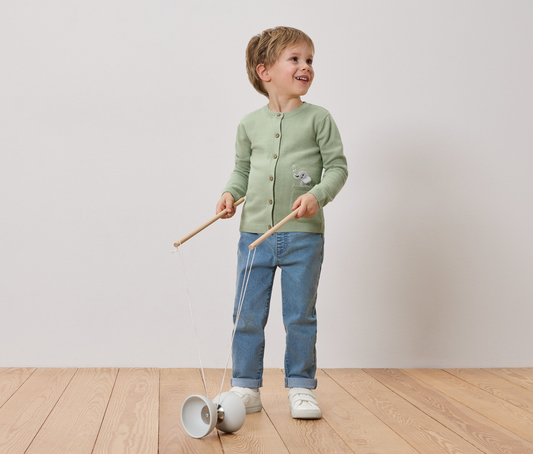 Ein lächelnder Junge mit blonden Haaren spielt mit einem Diabolo. Er trägt eine grüne Strickjacke mit Elefantenmotiv, Jeans und weiße Turnschuhe.