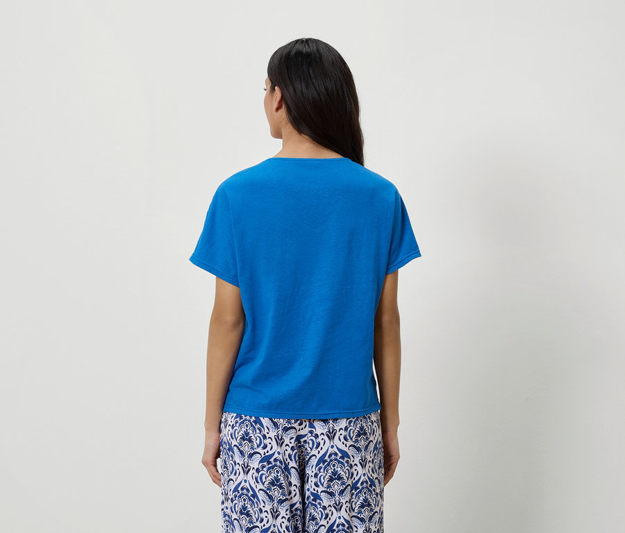 Rückansicht einer Frau mit dunklen Haaren, die ein blaues T-Shirt und eine blau-weiß gemusterte Hose trägt.
