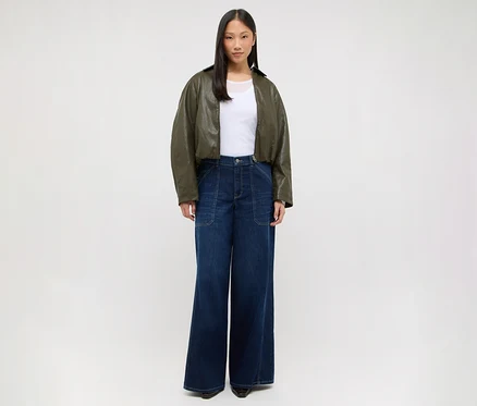 Eine Frau steht frontal, trägt ein weißes T-Shirt, eine kurze olivgrüne Jacke und eine dunkelblaue weite Jeans.