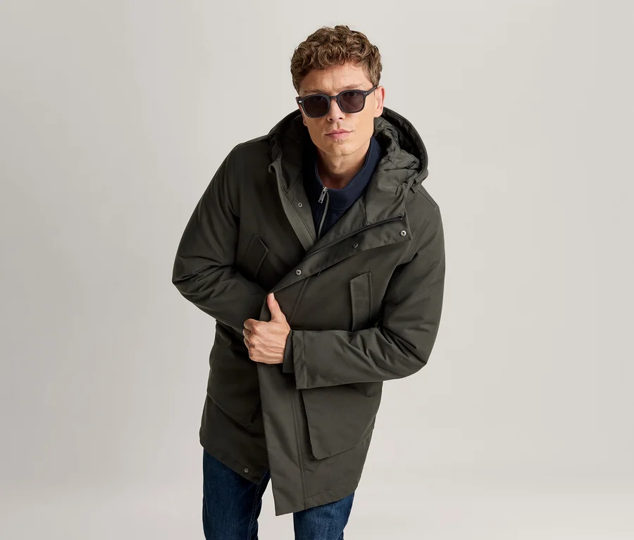 Mann in dunkelgrünem Kapuzenparka, blauem Reißverschlussoberteil und Jeans, trägt Sonnenbrille und hält seine Jacke.