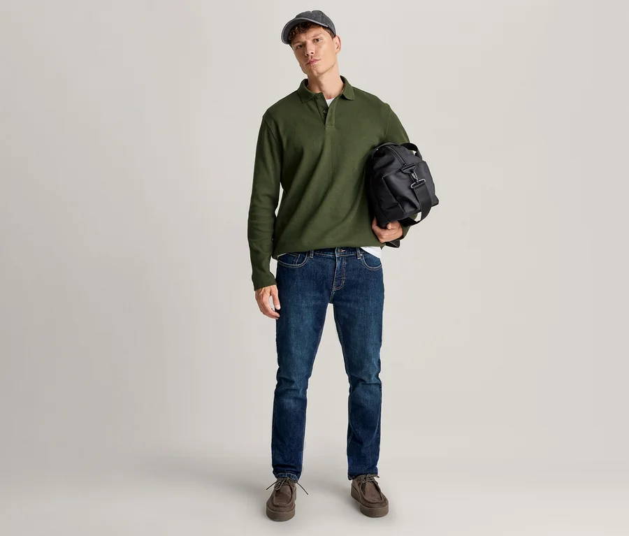 Ein Mann steht in einem grünen Langarm-Poloshirt, blauen Jeans, braunen Schuhen und einer dunklen Kappe und hält eine schwarze Reisetasche.