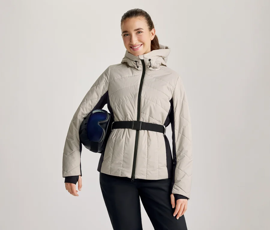 Lächelnde Frau in beiger Steppjacke mit schwarzen Seitenpartien und schwarzem Gürtel, schwarzer Hose, hält einen dunkelblauen Skihelm mit Skibrille unter dem linken Arm.