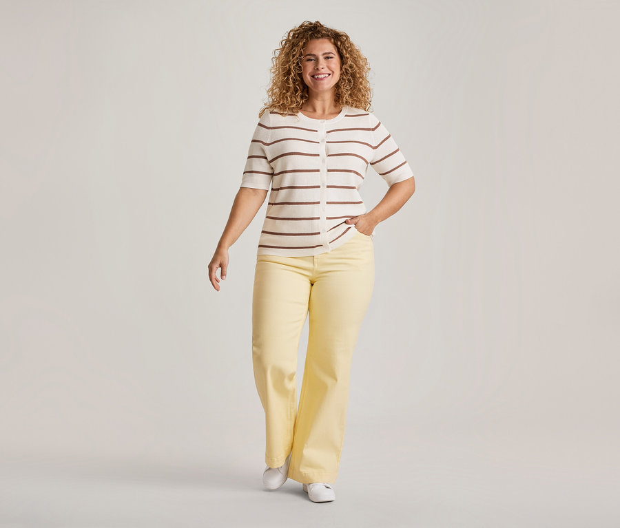 Eine lächelnde Frau mit lockigem Haar geht vorwärts, trägt einen cremefarbenen, kurzärmeligen Cardigan mit braunen Streifen, eine gelbe weite Hose und weiße Schuhe. Eine Hand steckt in ihrer Tasche.