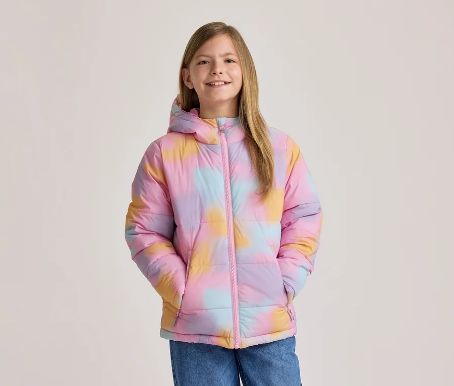 Lachendes Mädchen in bunter Tie-Dye-Pufferjacke in Rosa, Gelb und Blau mit Kapuze und rosa Reißverschluss, Hände in den Taschen.