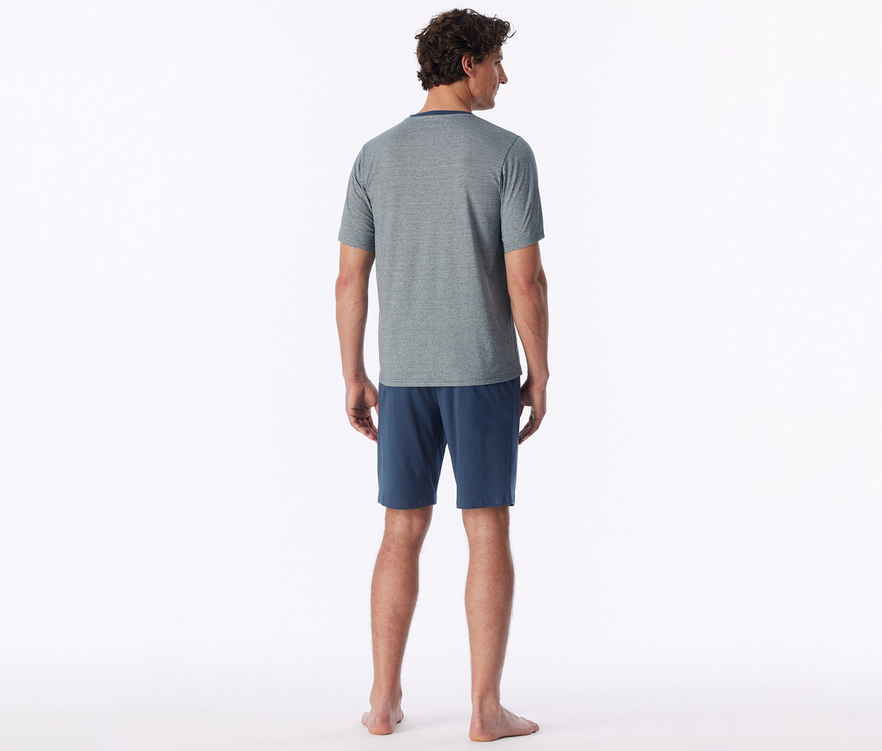 Mann von hinten, trägt ein graues T-Shirt und blaue Shorts, barfuß stehend.
