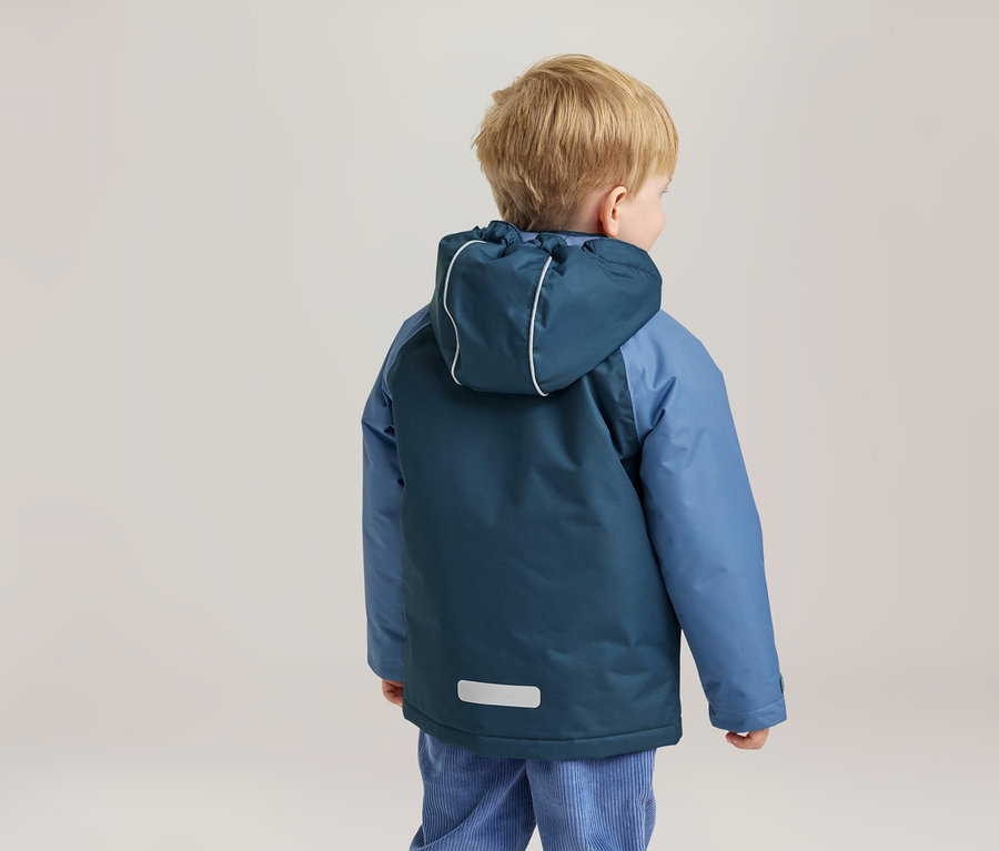 Kleines Kind von hinten gesehen, trägt eine zweifarbige blaue Kapuzenjacke mit reflektierenden Details und hellblaue Cordhosen. Das Kind hat blonde Haare und blickt nach rechts.