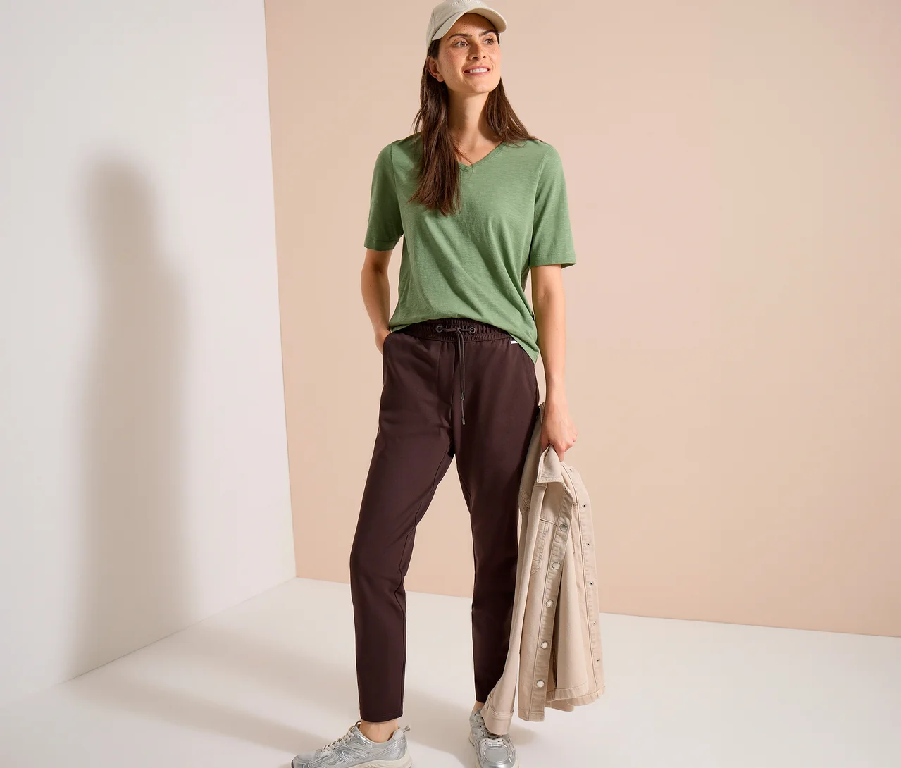 Eine Frau mit beiger Kappe trägt ein grünes V-Ausschnitt-Shirt, eine braune CECIL Jerseyhose und silberne Sneaker, hält eine beigefarbene Jacke.