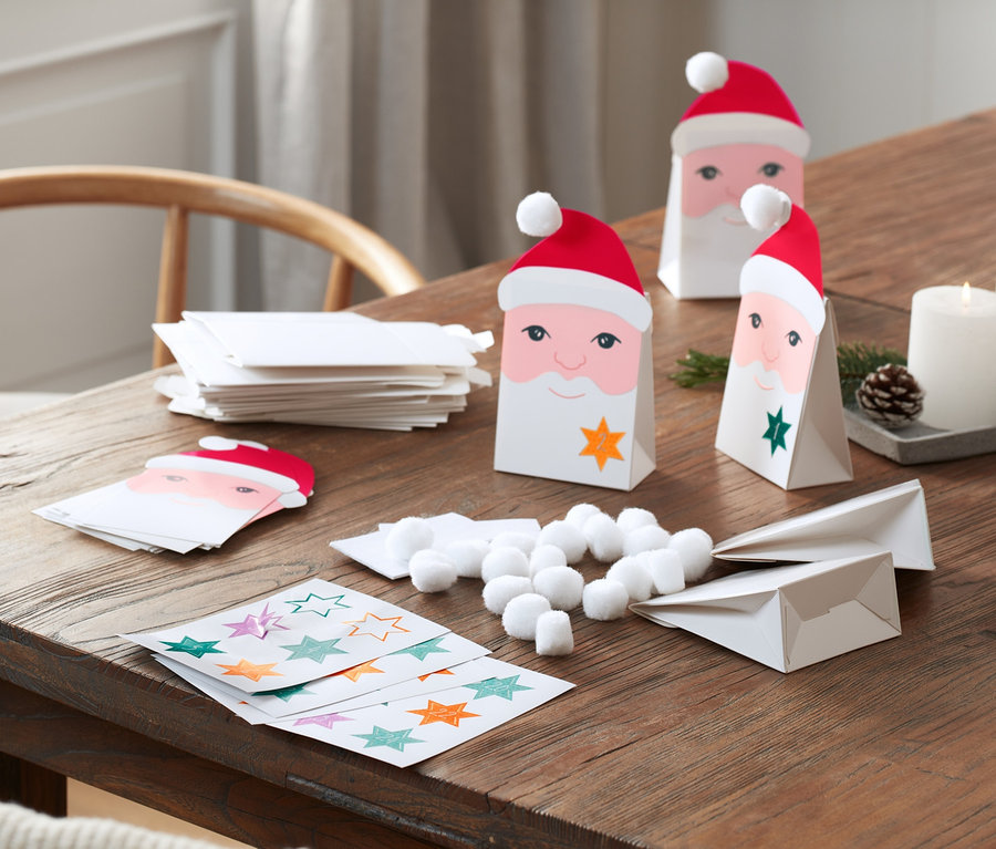 Auf einem Holztisch liegt ein DIY-Adventskalender-Set mit Santa-Gesicht-Papiertüten, Sternaufklebern, Wattebällchen und Kerzen.