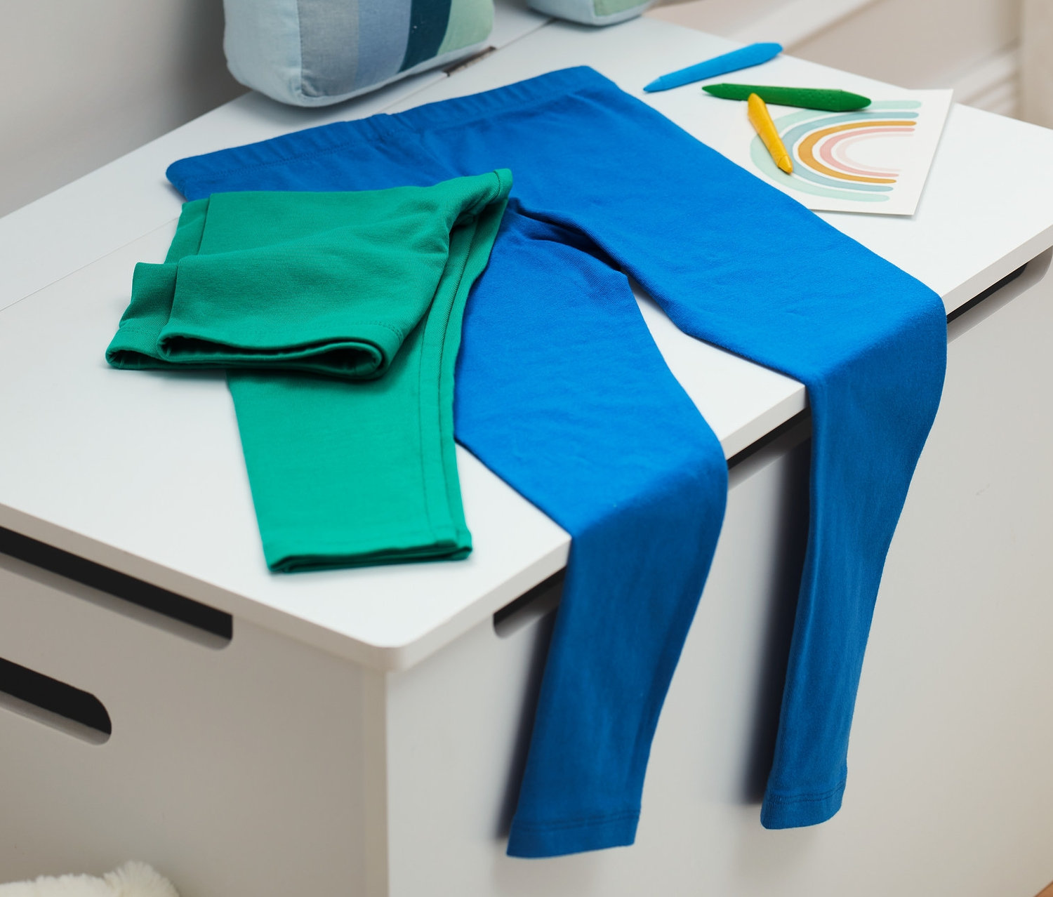 Auf einer weißen Truhe liegen blaue und grüne Leggings, daneben eine Regenbogen-Dekoration und Buntstifte.
