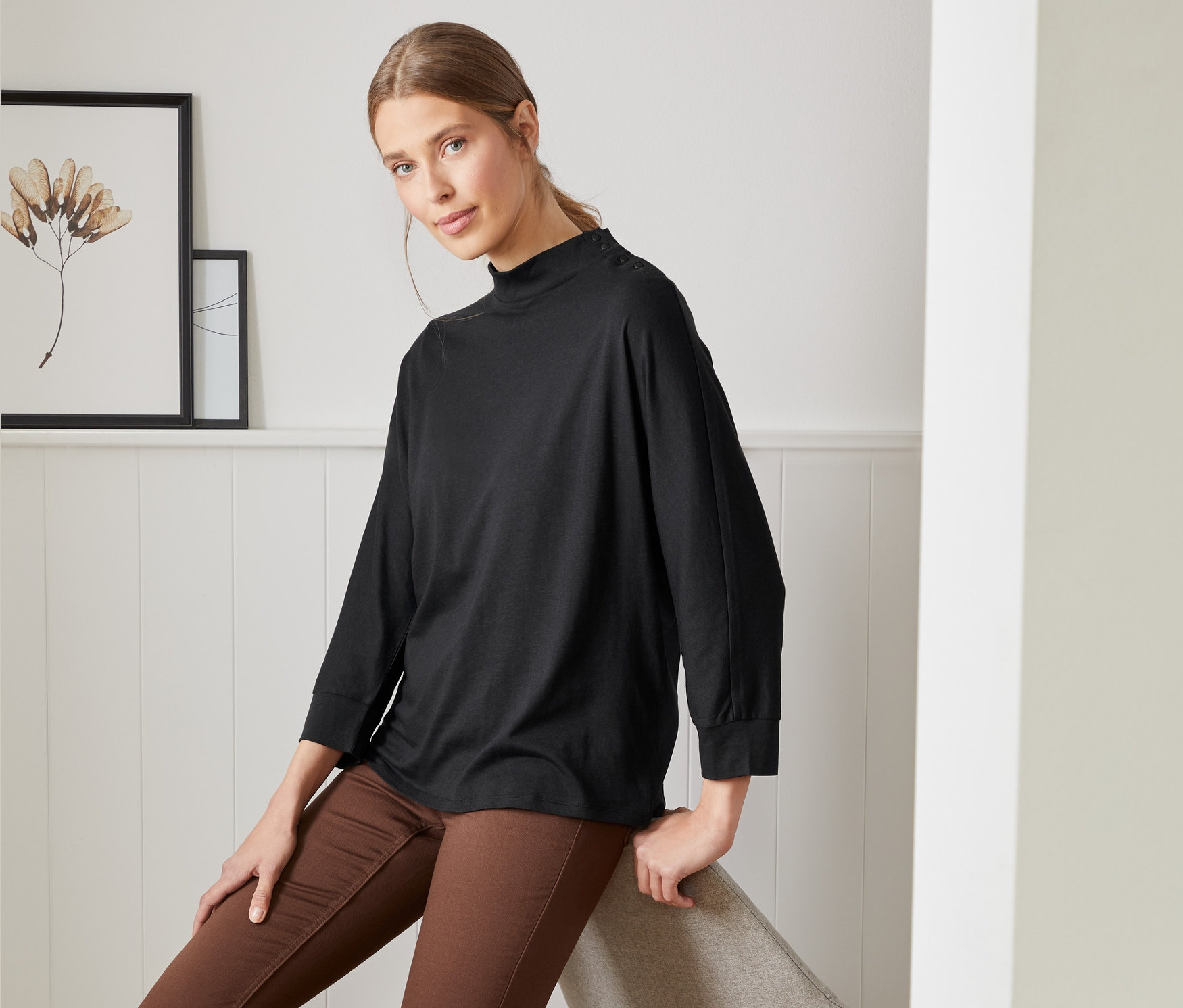 Frau sitzt auf einem Stuhl und trägt ein schwarzes Fledermausshirt mit Knöpfen und braune Hosen.