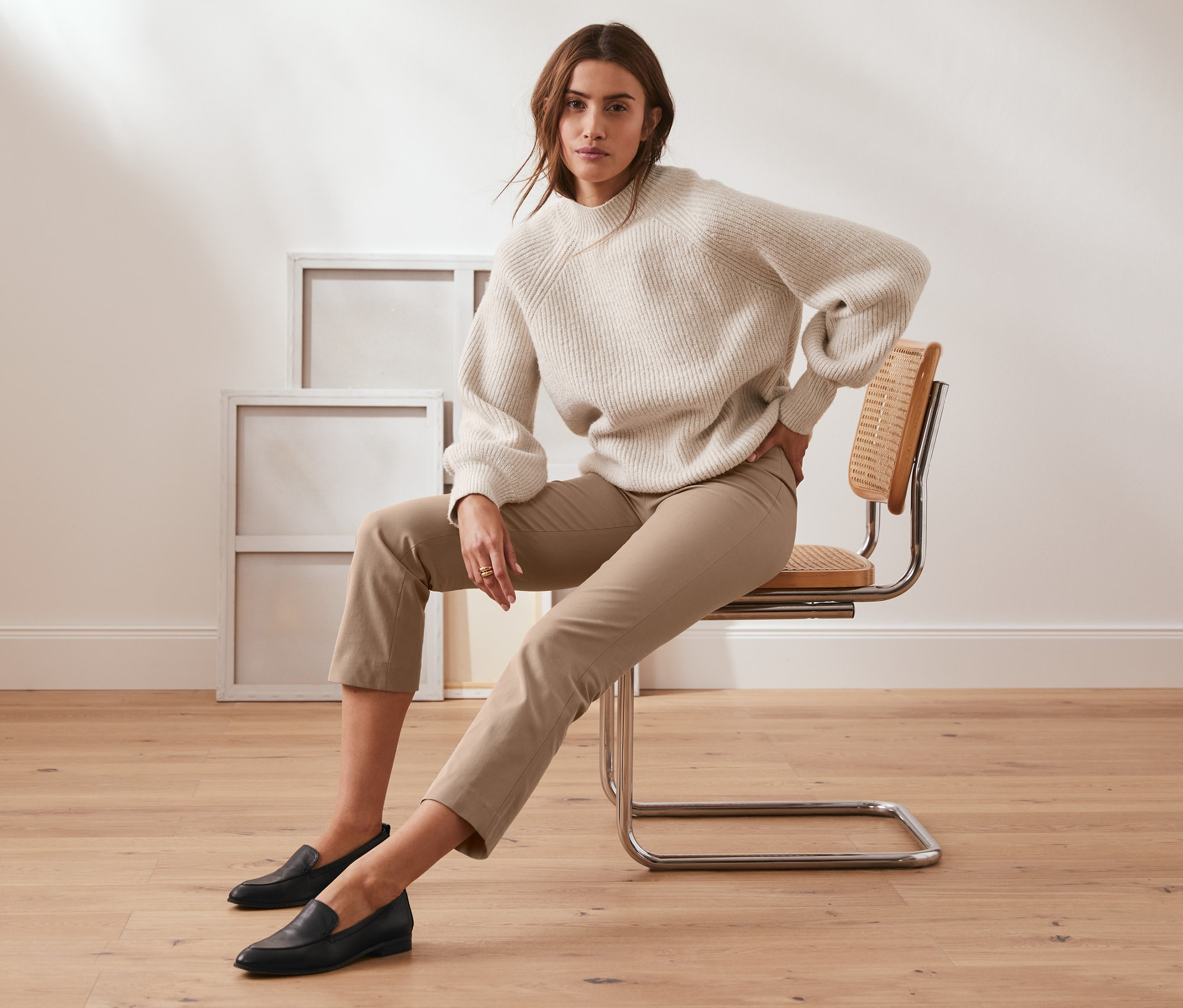 Frau sitzt auf einem Stuhl und trägt eine 7/8-Stretch-Hose in Camel und einen Pullover.