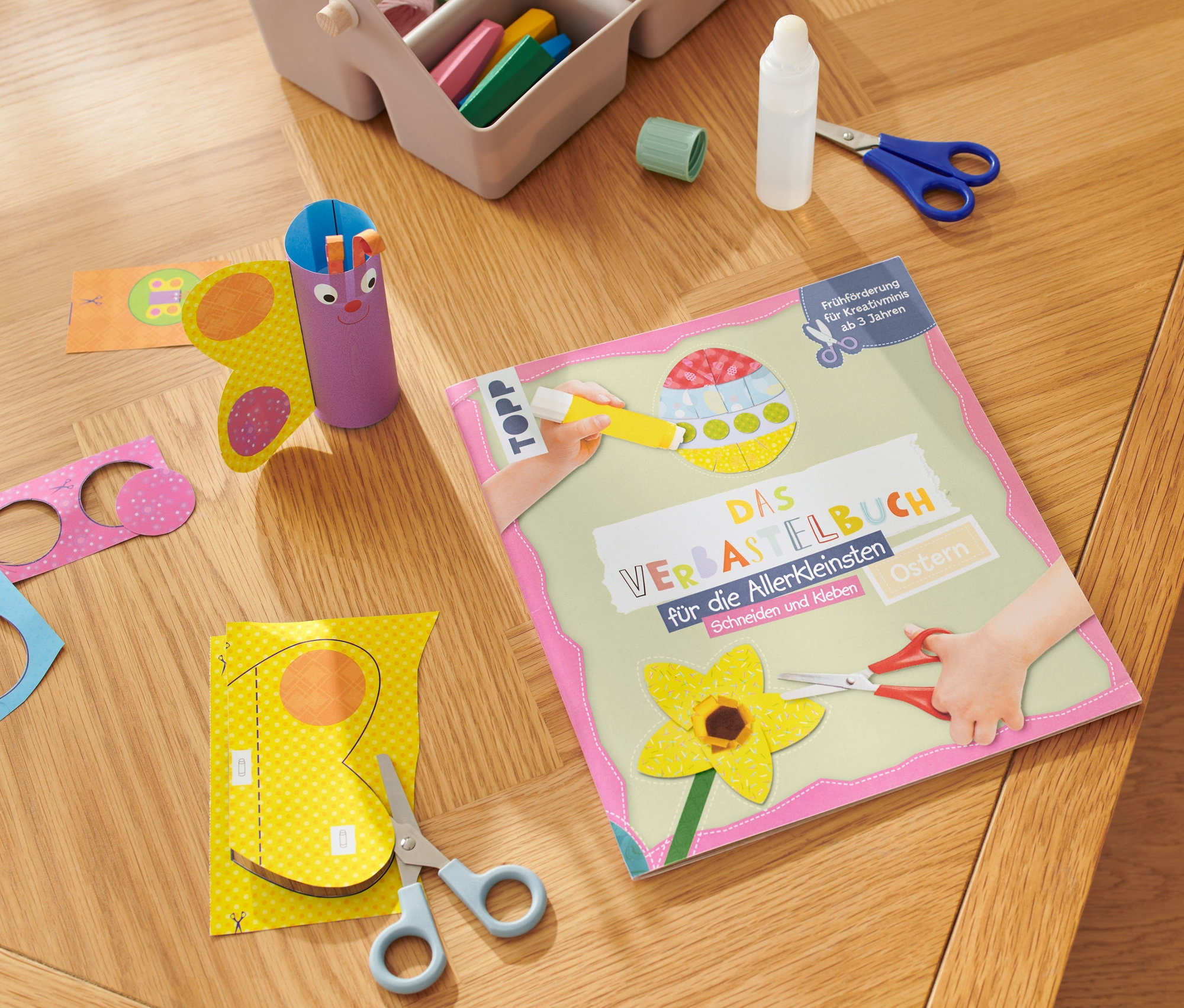 Auf einem Holztisch liegen das Buch "Das Verbastelbuch für die Allerkleinsten – Ostern", Scheren, Kleber und unfertige Papierarbeiten.