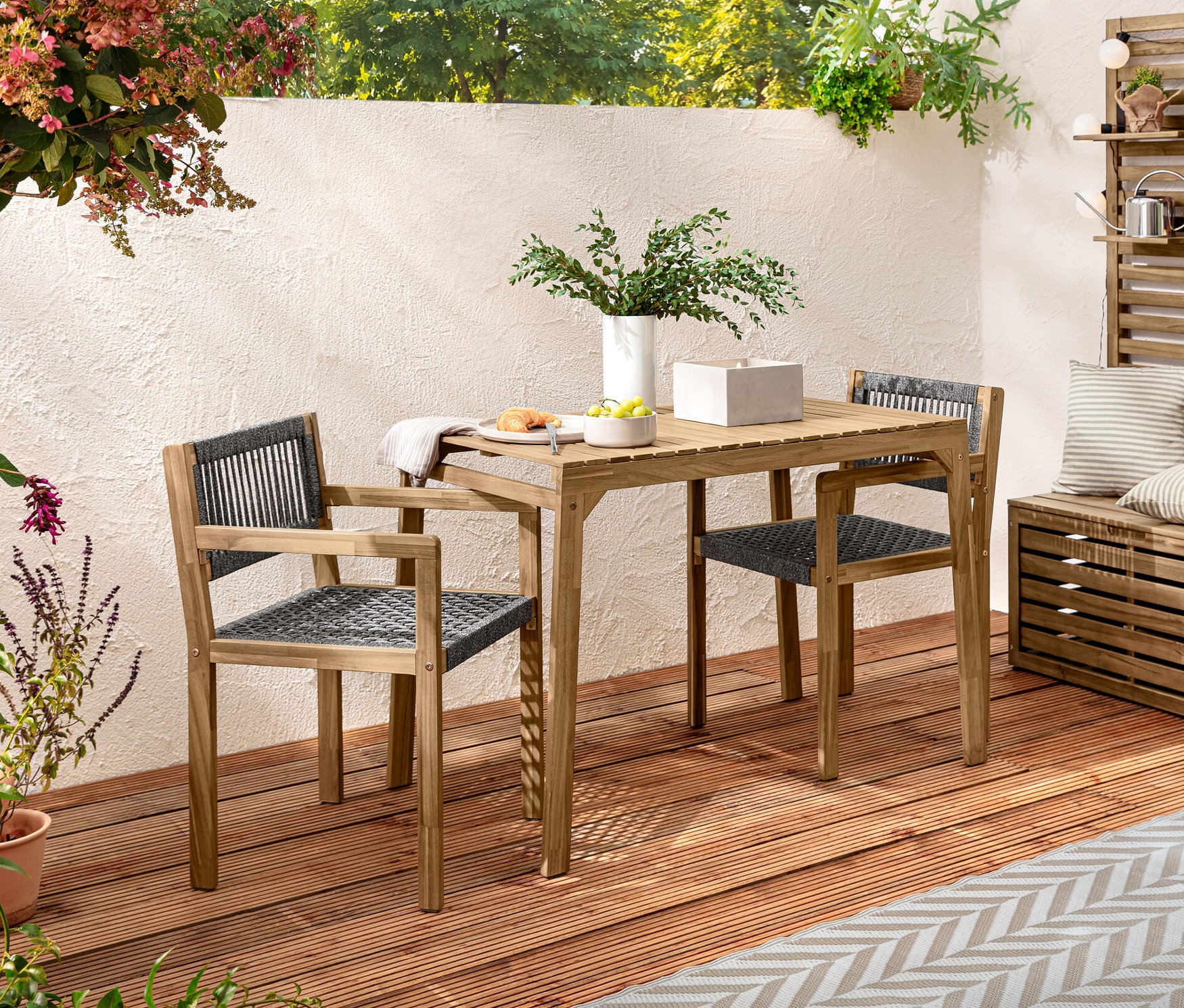 Ein Balkon-Set mit Textilgeflecht steht auf einer Holzterrasse. Auf dem Tisch steht eine XL-LED-Echtwachskerze im Betontopf. Dekoriert mit 2 Outdoor-Kissen in Leinen-Haptik und einer LED-Outdoor-Lichterkette.