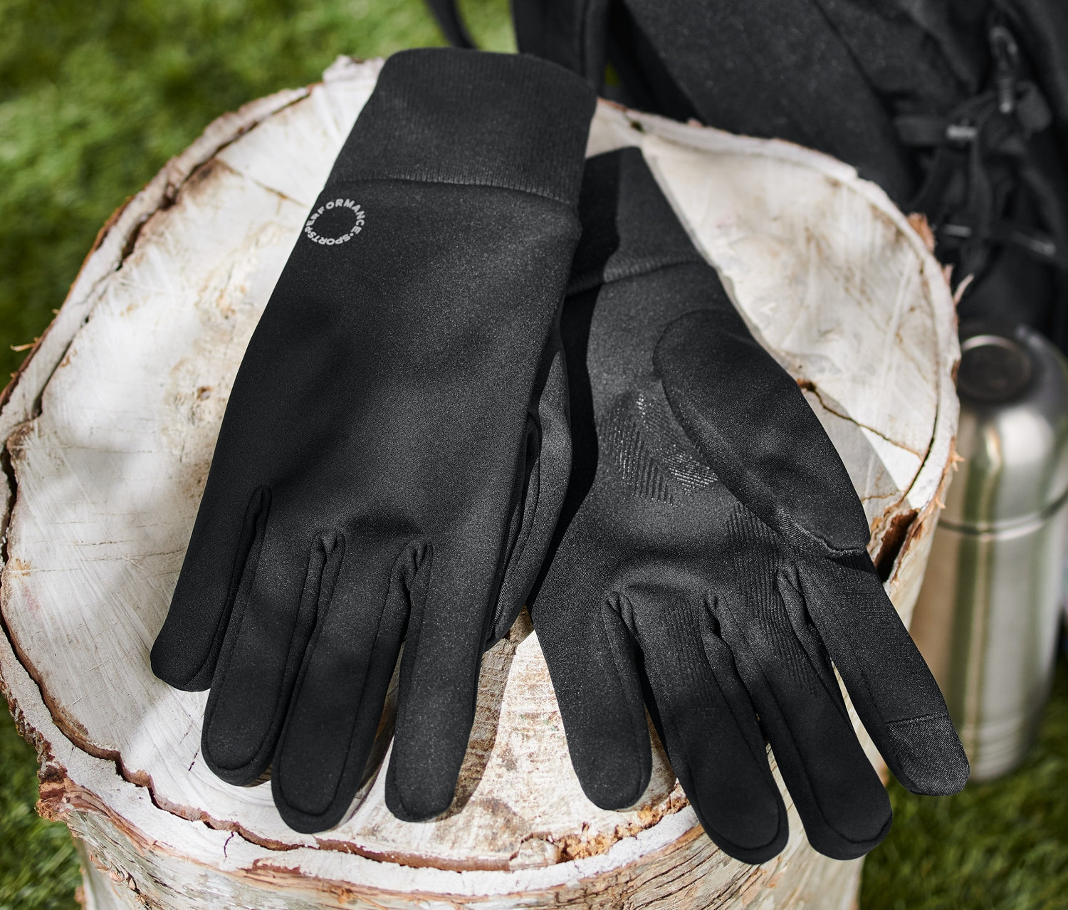 Schwarze Windprotection-Handschuhe liegen auf einem Baumstumpf neben einer Thermoskanne, einer Karte und einem Rucksack.