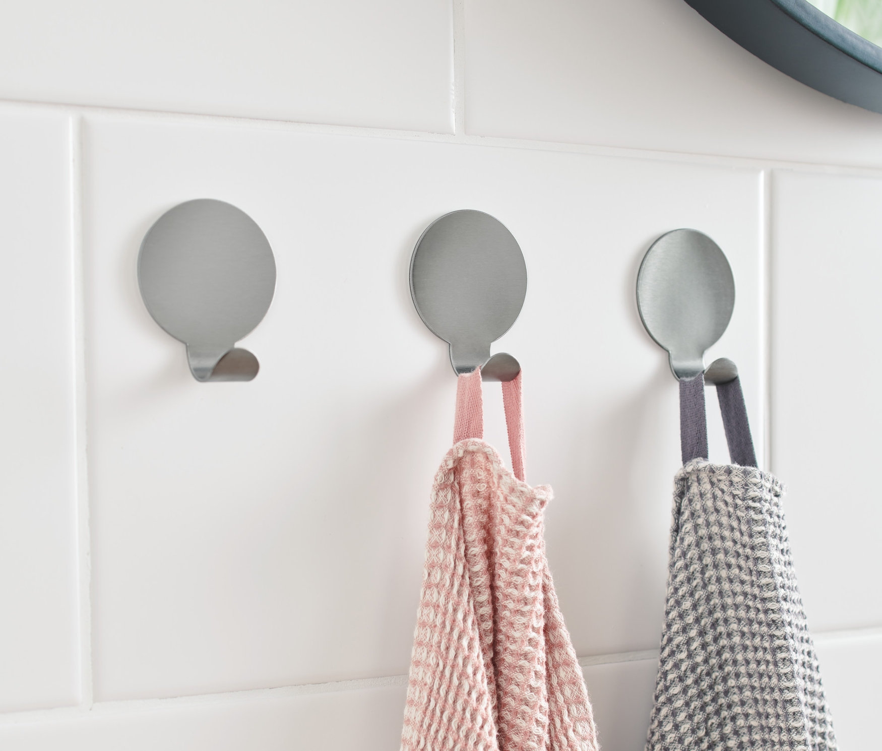 Drei silberne Haken sind an weißen Fliesen befestigt. An zwei davon hängen Handtücher.