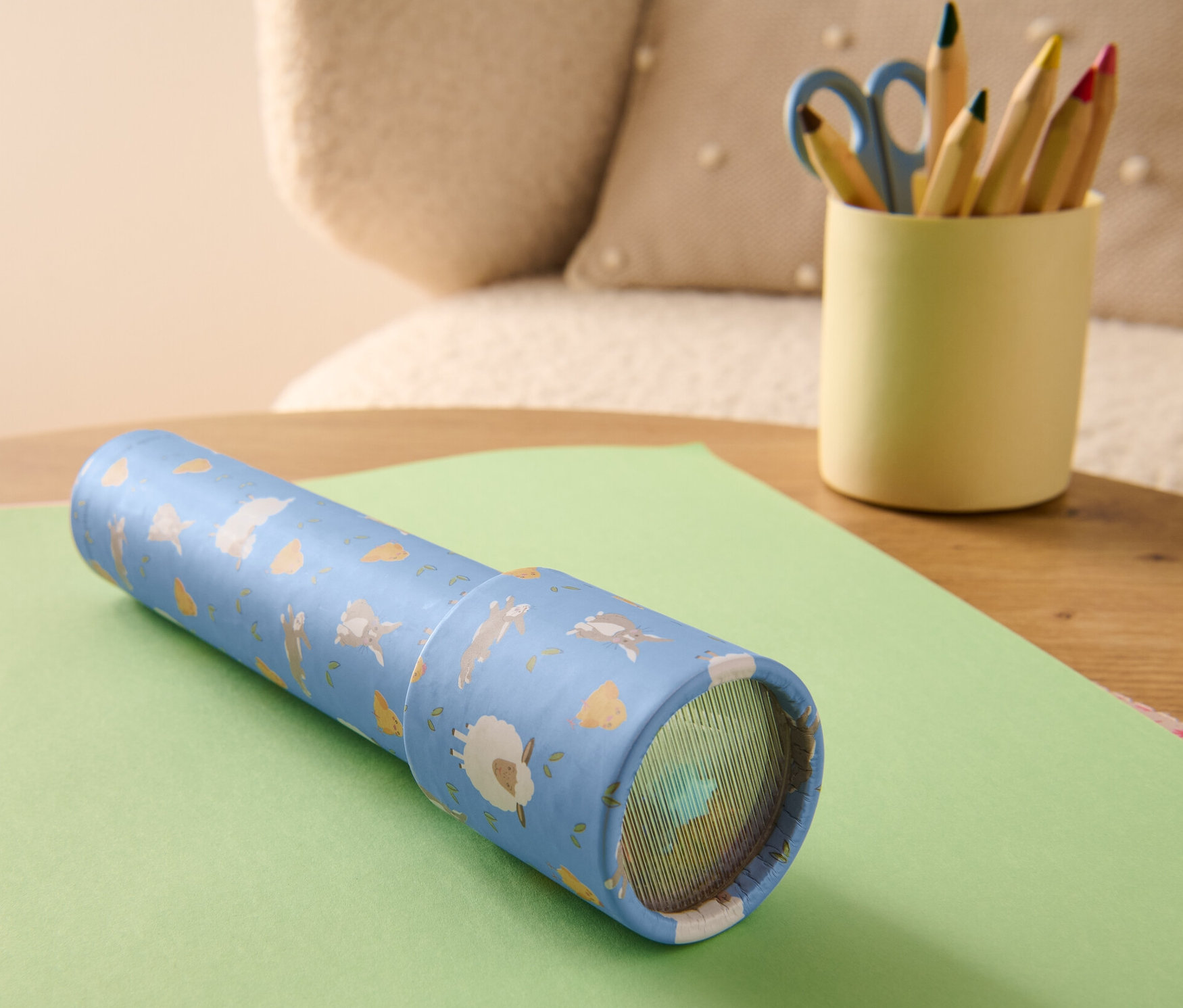 Ein Kaleidoskop liegt auf einem grünen Blatt Papier. Daneben steht ein Becher mit Buntstiften und einer Schere.