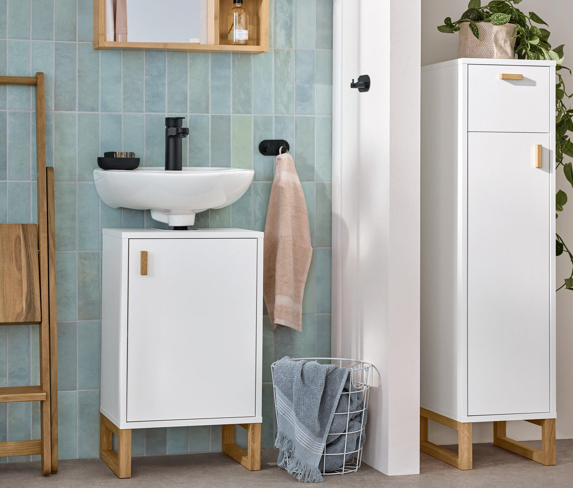 Ein Badezimmer zeigt einen kleinen Waschbeckenunterschrank mit Tür, einen schmalen Bad-Beistellschrank mit Schublade und Tür sowie einen Spiegel mit Ablageflächen.