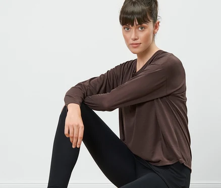 Frau sitzt auf einem Holzhocker und trägt ein braunes 2-in-1-Sportshirt und schwarze Leggings.