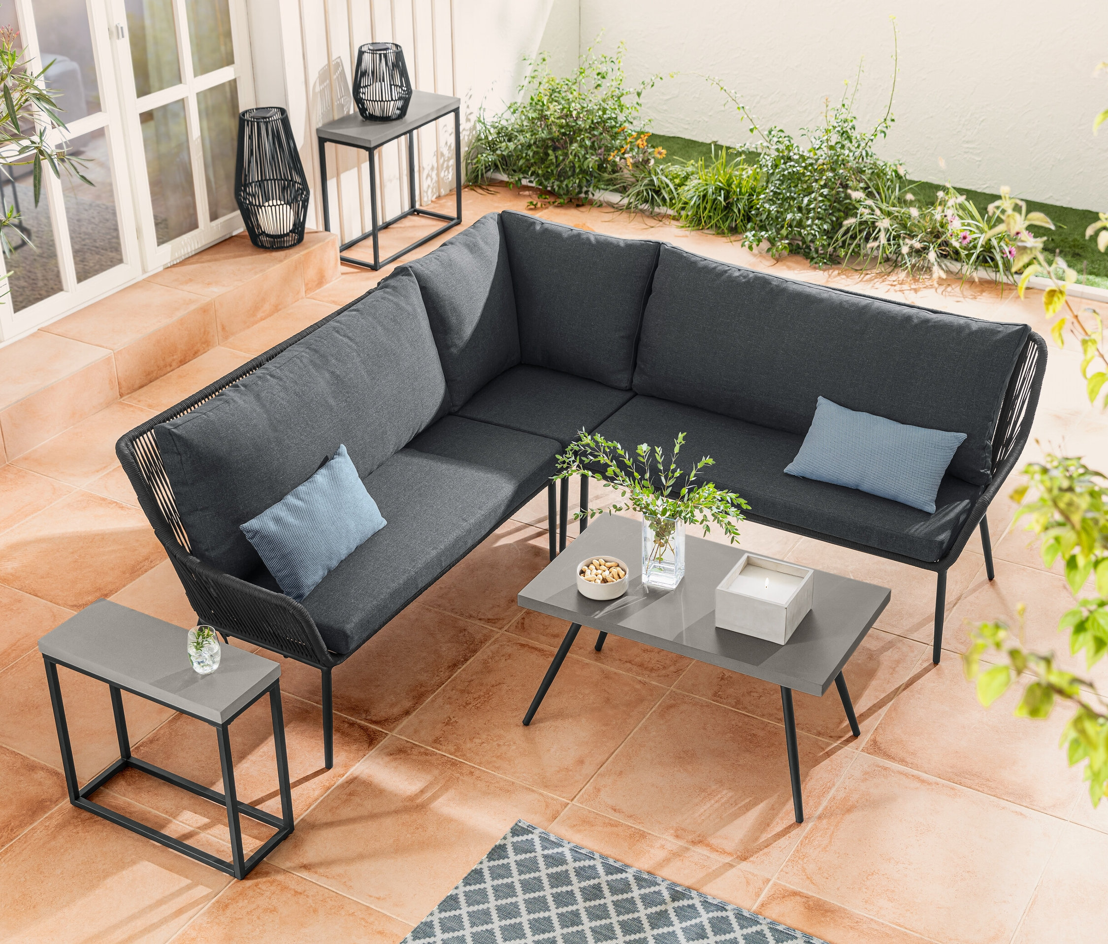 Eine Lounge-Ecke mit Textilgeflecht steht neben einem Couchtisch mit Polyzement-Tischplatte. Ein Konsolentisch-Set mit Polyzement-Tischplatte steht neben einer Solar-Laterne mit Textilband.