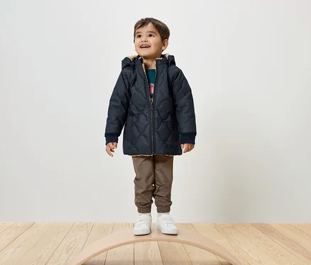 Ein Junge steht auf einem Balance Board und trägt eine marineblaue Steppjacke, braune Hosen und weiße Turnschuhe.