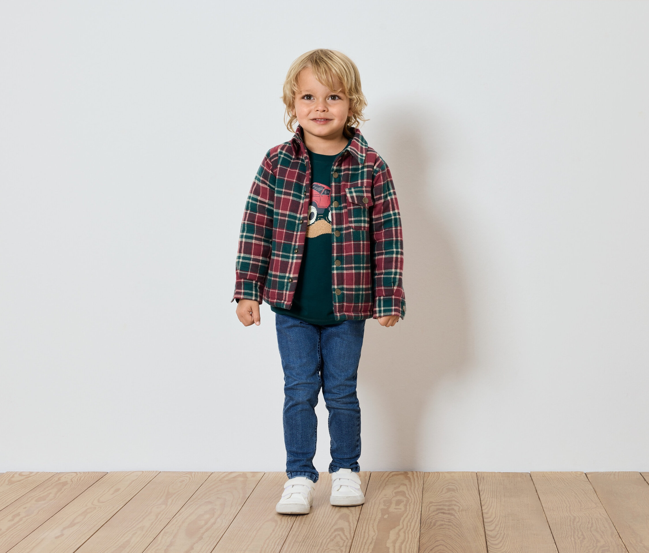 Blonder Junge steht vor einer weißen Wand, trägt ein grünes T-Shirt, ein kariertes Hemd, Jeans und Turnschuhe.