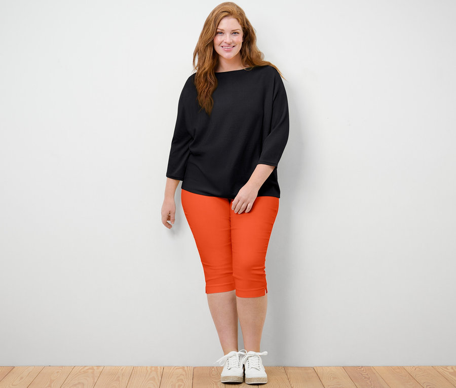 Frau steht und trägt eine orangefarbene 3/4-Stretchhose und einen schwarzen Feinstrickpullover.