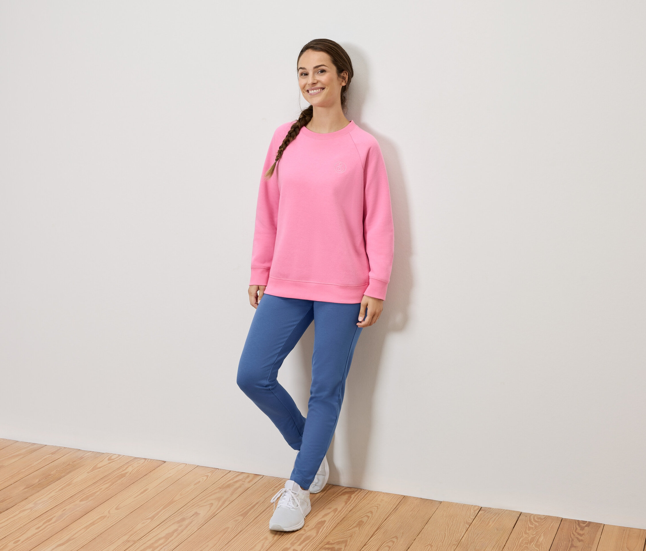 Frau mit geflochtenem Haar in rosa Sweatshirt, blauen Hosen und weißen Schuhen, die sich an eine Wand lehnt.