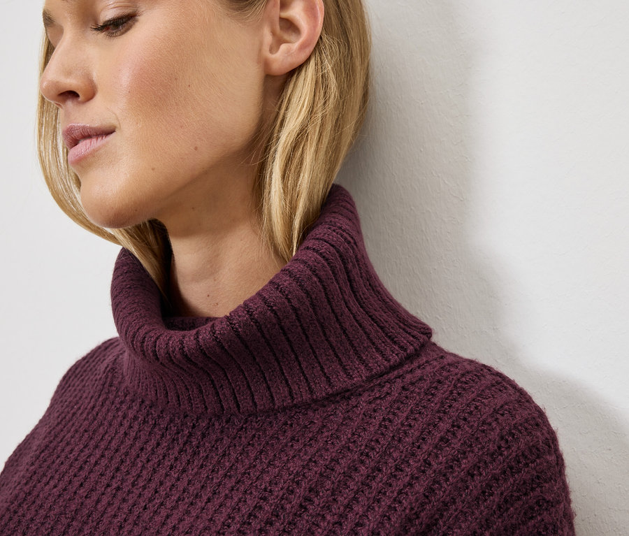 Porträt einer blonden Frau mit einem bordeauxroten Strickrollkragenpullover.