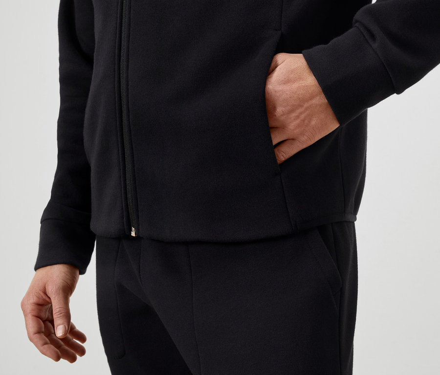 Mann hat Hand in der Tasche einer schwarzen Reißverschlussjacke und trägt eine schwarze Hose.