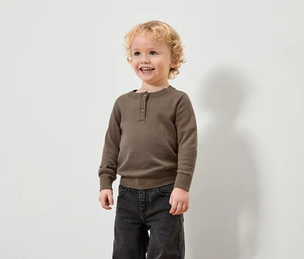 Ein Junge mit blonden Locken trägt einen braunen Pullover und schwarze Jeans.