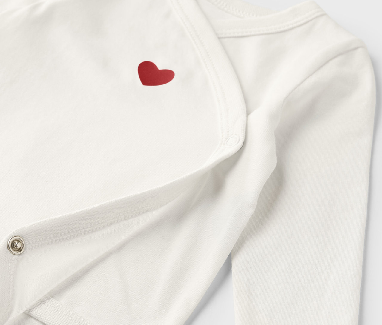 Detailaufnahme eines weißen Baby-Bodys mit Mitwachsfunktion und rotem Herz.