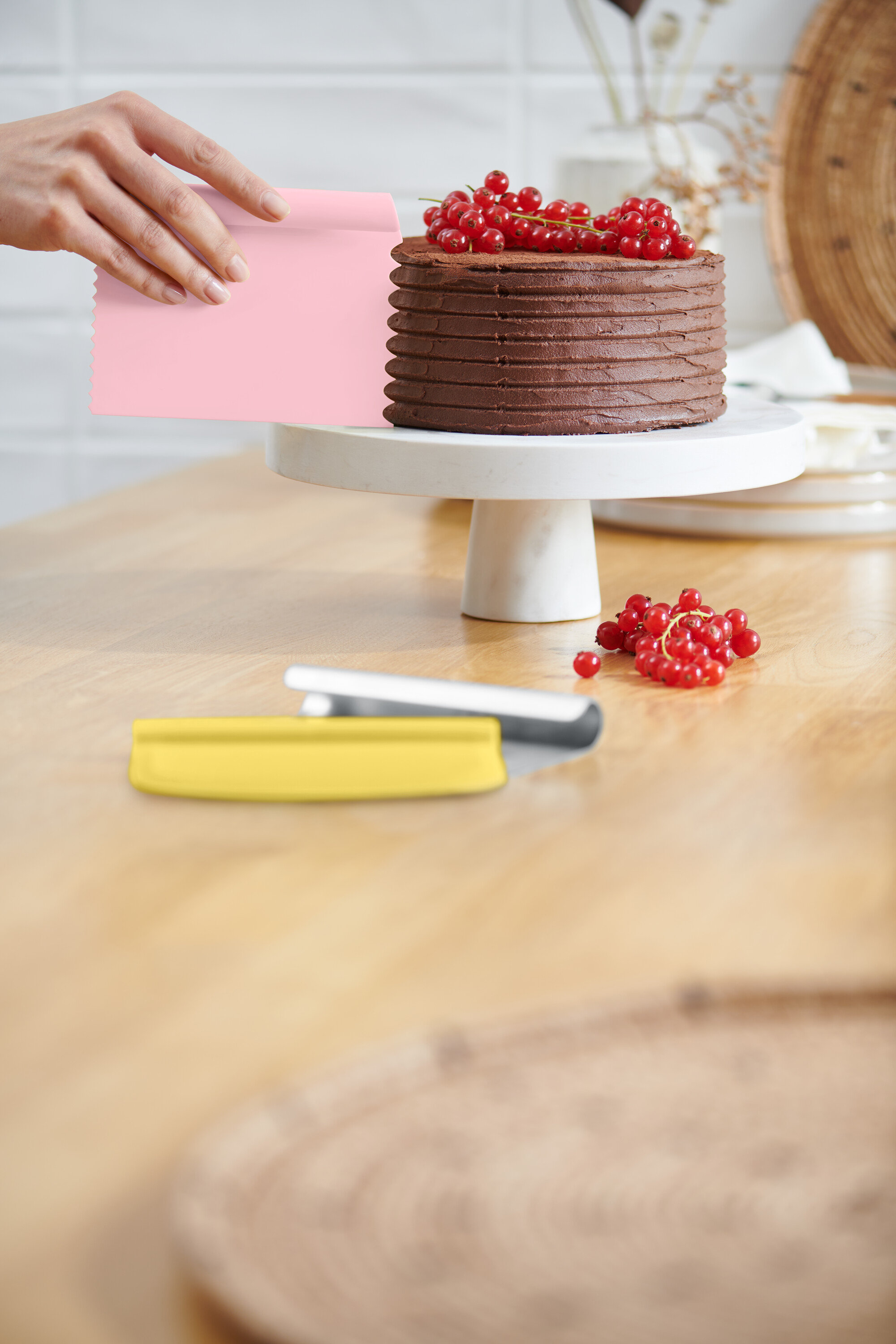 Eine Hand glättet die Glasur einer Schokoladentorte mit einem rosa Teigschaber. Die Torte steht auf einem weißen Tortenständer und ist mit roten Johannisbeeren belegt. Ein gelber Teigschaber liegt auf dem Tisch.