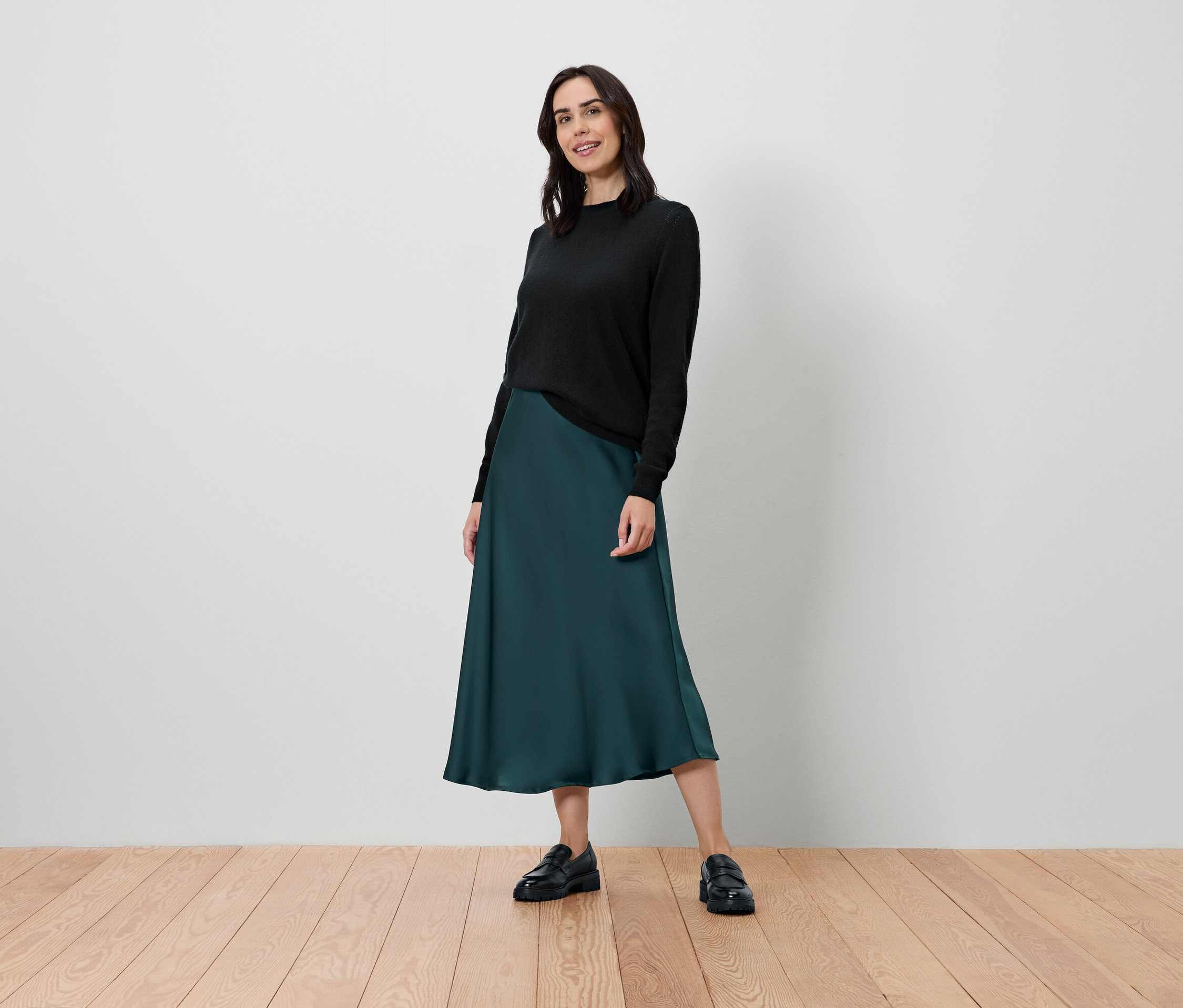 Eine Frau steht und posiert in einem schwarzen Pullover und einem dunkelgrünen Rock.