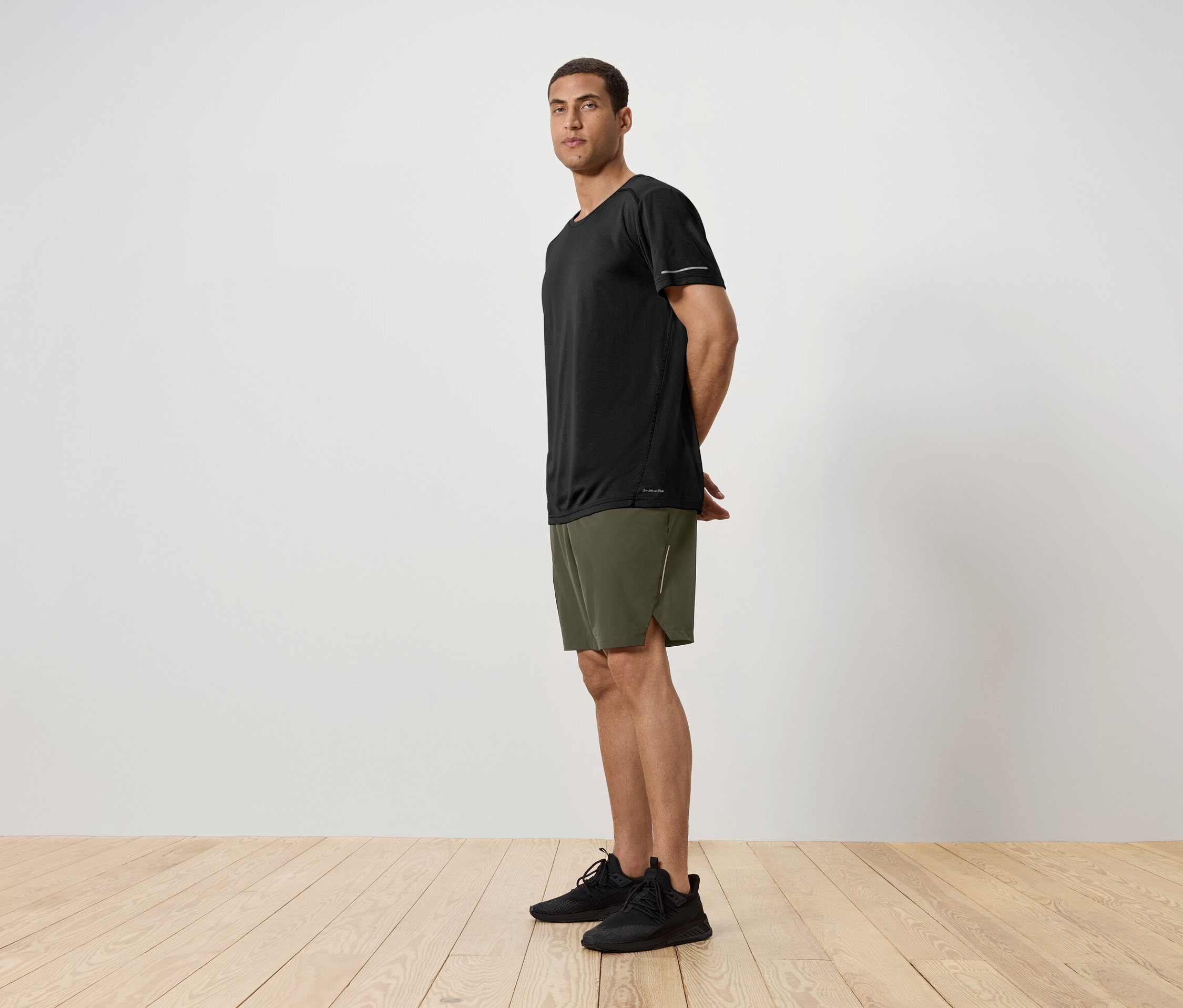 Seitenansicht eines Mannes in einem schwarzen T-Shirt, grünen Shorts und schwarzen Turnschuhen, der auf einem Holzboden vor einer weißen Wand steht.